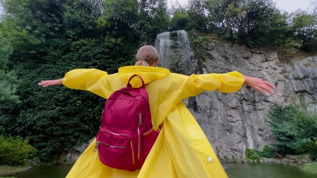 Woman wearing a yellow raincoat and backpack raising arms in front of a rocky waterfall in a green park in Trabzon, expressing freedom, energy and outdoor adventure
