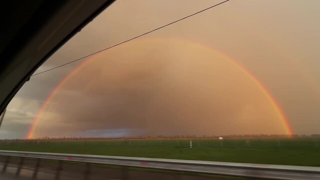 View from moving car through side window showing double rainbow over countryside landscape. Scenic roadside with guardrail and green fields under dramatic sky. Concept of travel, weather and journey