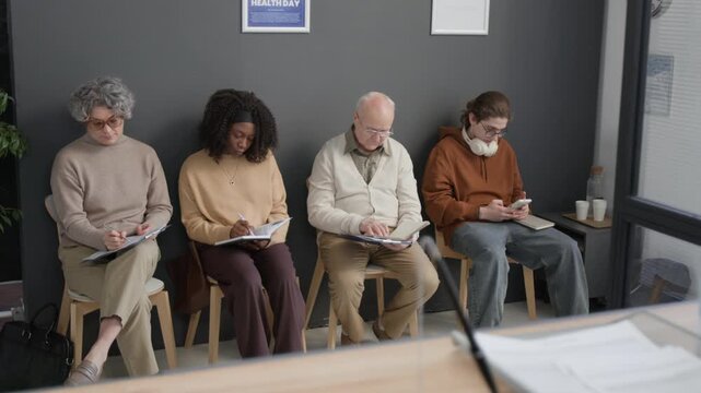 Diverse men and women sitting on chairs in waiting hall in front of hospital reception desk, signing agreement, making notes and using smartphone