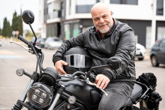 Happy middle aged man sitting on classic motorcycle holding helmet, enjoying urban adventure and freedom ride