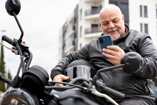 Middle aged man biker engaging with smartphone on motorcycle, checking messages, navigating urban surroundings