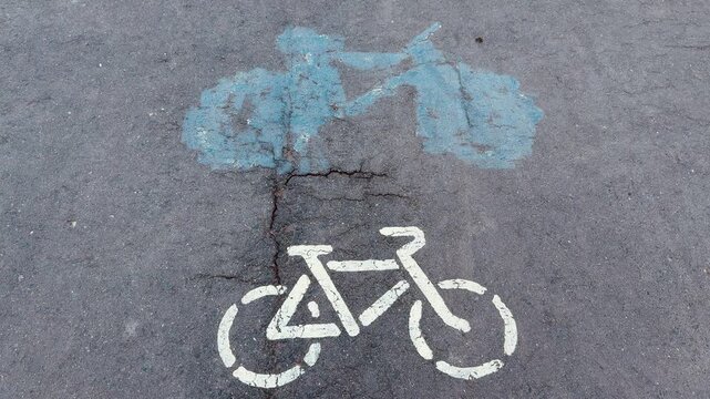 Faded bicycle lane stencil on asphalt, white bike symbol beneath old blue smudge, worn tarmac texture captured in closeup top down.