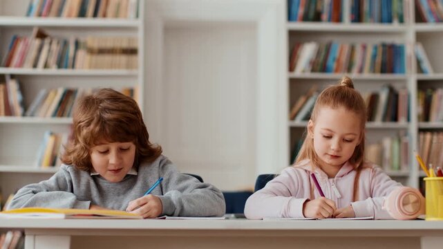 Two children are sitting at a table in a bright classroom with bookshelves filled with books. They hold writing tools and appear focused on their work. The atmosphere is engaging for learning.