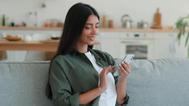 Happy relaxed Indian woman using mobile phone at home browsing social media chatting smartphone scrolling online shopping Arabian female ethnic girl holding cellphone gadget addict technology indoors