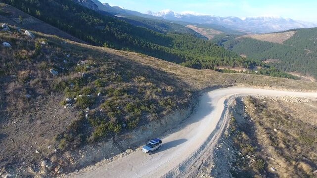 Aerial view of car driving along winding mountain road toward green forest valley. Vehicle follows curving route through rugged terrain into dense woodland landscape.