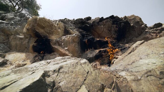 Burning methane flame emerges from rock crack as natural gas leaks from underground earth surface. Combustible seep creates fiery vent between stones showing geological energy release.