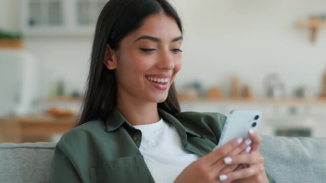 Excited joyful Indian woman at home using mobile phone celebrating win victory success smiling looking at camera shopping online discount Arabian female girl champion positive cellphone game indoors