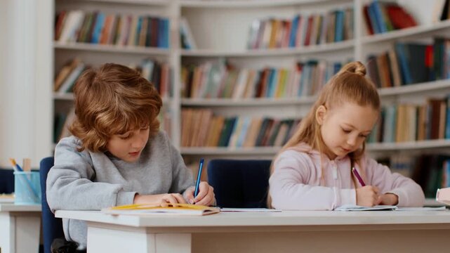 Two children focus on their schoolwork at a white table in a classroom. A boy has curly hair and wears a light sweater. A girl tied her hair back and writes with a pen.