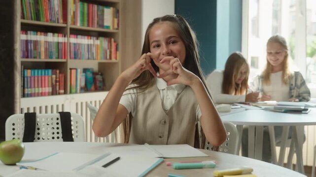 Smiling Caucasian little girl child making heart gesture with fingers in classroom happy glad kid schoolgirl school student affection cheerful pupil love symbol teenager enjoying lesson indoors class