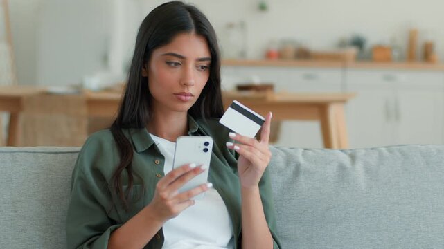 Serious Indian woman at home using phone making payment credit bank card mobile online shopping browsing Arabian female girl client buyer lady purchase pay order booking problem error debt indoors