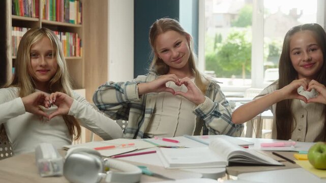 Joyful Caucasian three little girls children showing heart gesture with hands indoor class love friendship kids schoolgirls students teenagers smile looking camera education school teamwork together