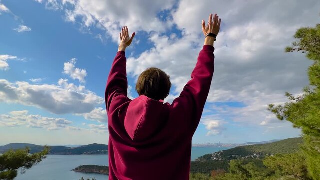 Back view of man raising arms toward cloudy sky while standing above sea and green hills near Istanbul. Freedom, travel and outdoor lifestyle concept