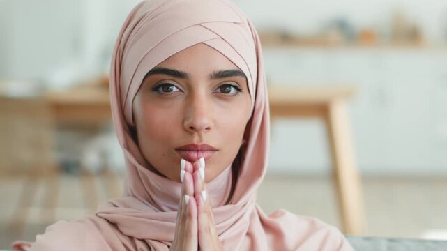 Serious concentrated Indian Muslim woman wearing pink hijab praying looking at camera at home indoors hopeful with folded hands spiritual Arabian Islam female peaceful Islamic girl lady faith devotion