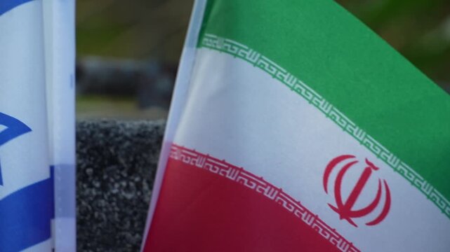 Miniature flags of Israel and Iran wave on flagpoles in an outdoor setting.