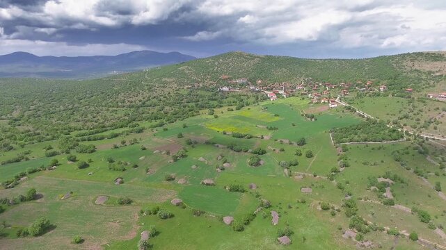Aerial Balkan mountain village homes beside forest, bright green fields, cloudy horizon. Overhead panorama reveals rural hamlet rooftops near woodland farms, patchwork meadows, distant ridgeline.