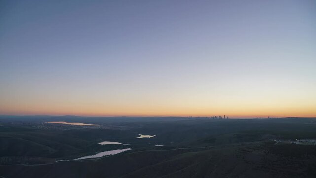 Timelapse sunset fades into night as city lights sparkle beside a winding lake and river. Afterglow turns to darkness under clear sky, showing urban illumination, still reservoir, far ridgelines.