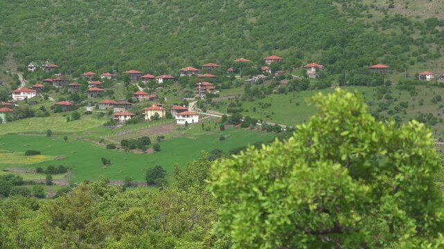 Balkan mountain village houses near forest and green fields, wide rural panorama view. Scenic hillside hamlet rooftops amid woodland farms, lush meadows, distant ridges under haze.