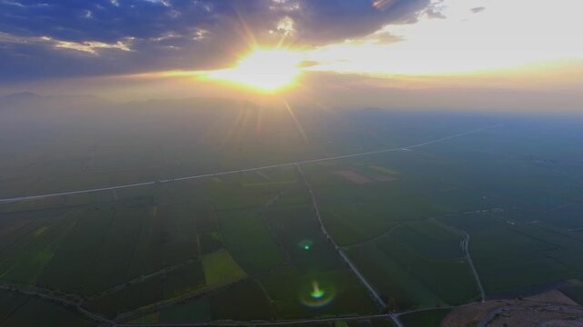 Aerial drone view captures sunset spreading warm light across farmland on a broad flat plain. Rural fields and evening colors create calm agricultural scenery above quiet cropland and farm plots.