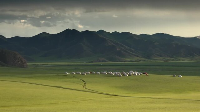 Mongolian ger village spreads across open steppe grassland beneath dark mountain ridge. Crowded yurt settlement lines the green plain, forming a nomad campsite near distant peaks.