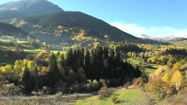Alps village in mountain valley with autumn forest, green meadows, panoramic cinematic landscape. Scenic hamlet among fall woods and rolling highlands, fairy tale countryside vista, vivid.