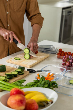 Chef's knife is slicing cucumber on wooden cutting board on white countertop, surrounded by produce