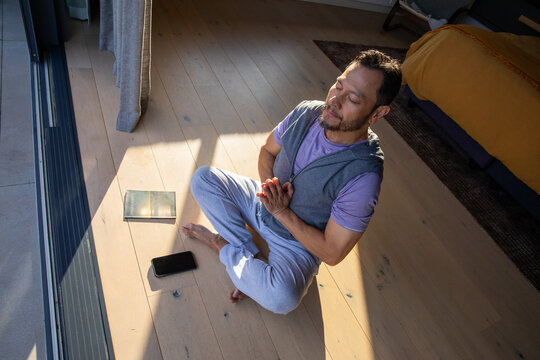 Middle-aged Asian man in purple shirt sitting cross-legged on floor near glass door, phone and book