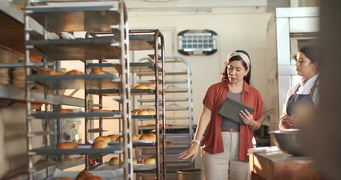 Hispanic owner coaching African American baker in bakery, entering checking racks and using tablet