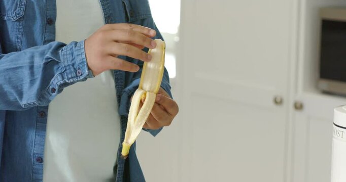 African American man holding partly peeled banana pinching peel and pulling making snack in kitchen