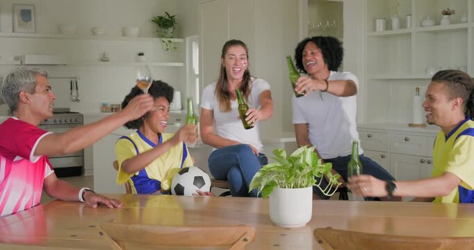 Diverse friends in jerseys toasting bottles after goal holding soccer ball at home kitchen table