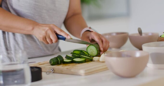 Placing cucumber, woman slicing even rounds with blue-handled knife, holding steady, prepping salad