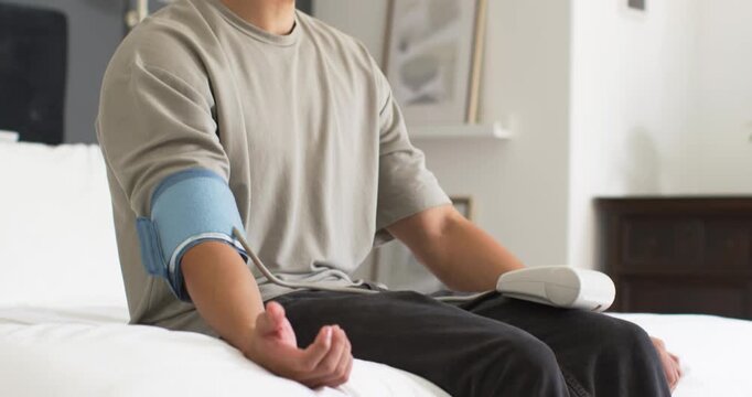 Asian man sitting on bed, wearing grey T-shirt and blue cuff, reading monitor displaying results