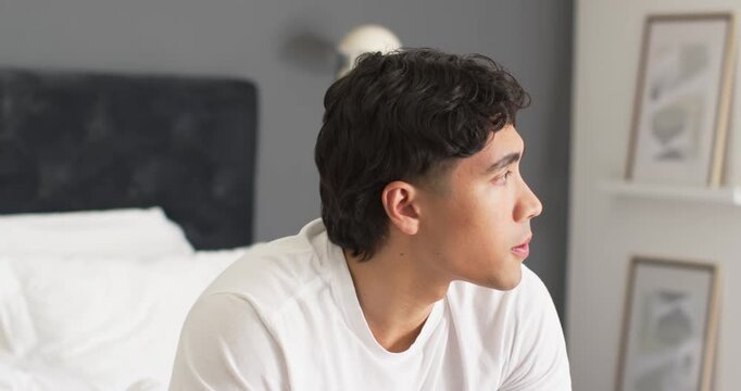 Asian man sitting on bed edge in bedroom, turning toward framed prints, wearing white T-shirt