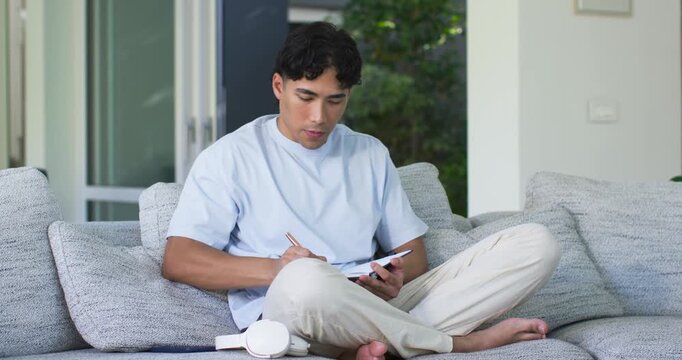 Asian adult male sitting on gray sofa writing planner while checking smartphone, headphones resting