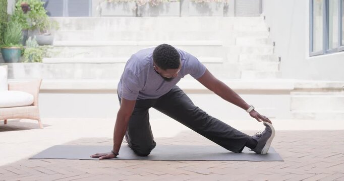Man wearing t-shirt kneeling on mat on brick patio warming up before workout reaching shoe