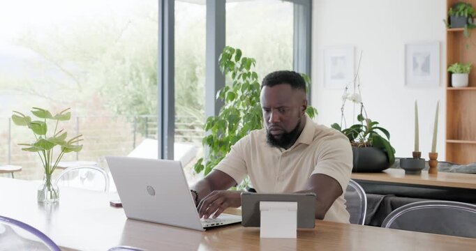 Remote worker typing on laptop, tapping tablet on stand, reviewing content, resolving business task