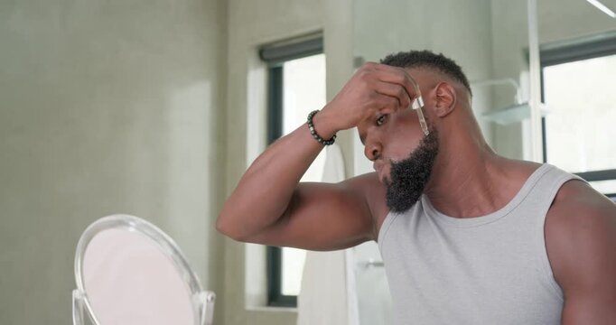 Man is picking dropper dispensing serum on brow, massaging beard for grooming while checking mirror