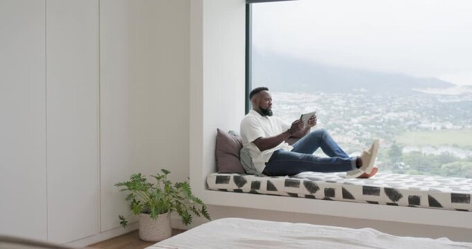 Vertical video: African man entering room with tablet sitting bay seat reading, resting, copy space