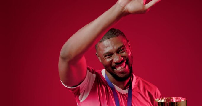 Mid-30s man wearing red sports jersey, holding gold trophy and medal, celebrating victory in studio