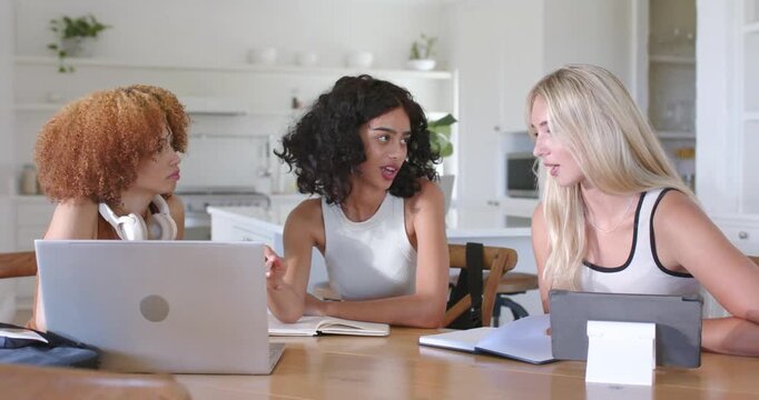 Three adult women sitting at kitchen table, central woman pointing at laptop, writing notes on task