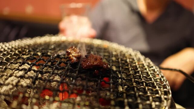 Close up of small pieces of meat grilling over hot coals on a metal grate with smoke rising.