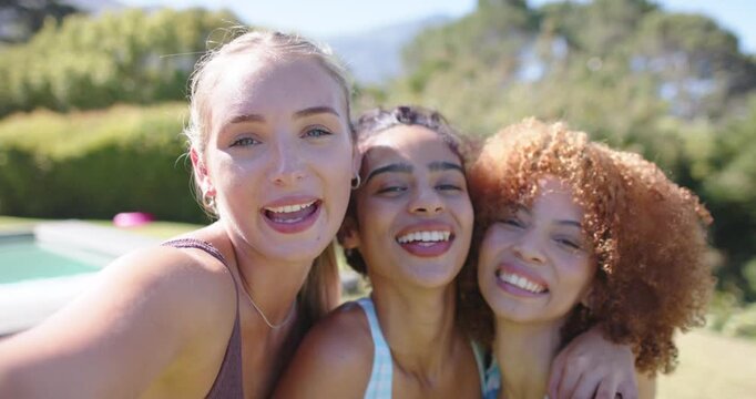 Leftmost woman is extending phone as three women are posing, laughing, and taking selfie poolside