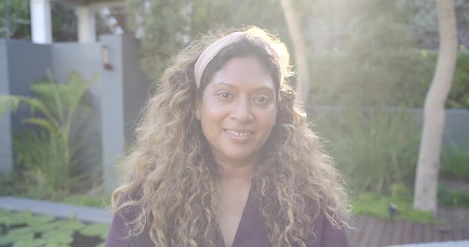 Indian woman in dark purple top with light headband squinting from sun then smiling near pond