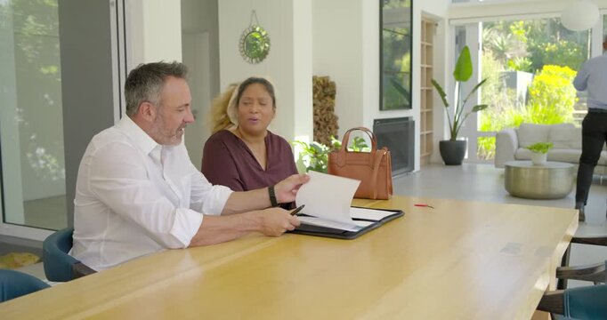 Diverse couple man opening folio and woman reading papers at dining table, man tapping pen agreeing