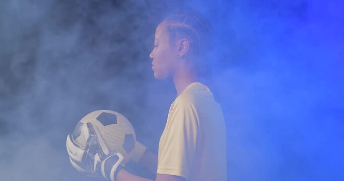 African American female keeper adjusting soccer ball grip with gloves as blue fog rolling in studio