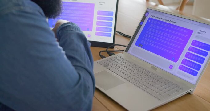 Adult African man reviewing purple chat on two-screen desk, tapping monitor and scrolling laptop