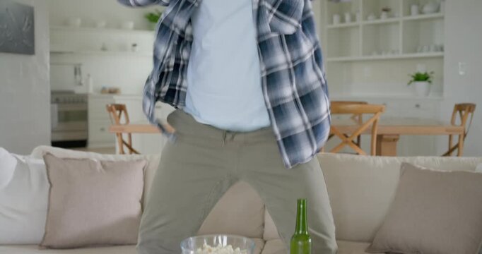 Adult African American man watching offscreen on couch, snacking popcorn, cheering, drinking bottle