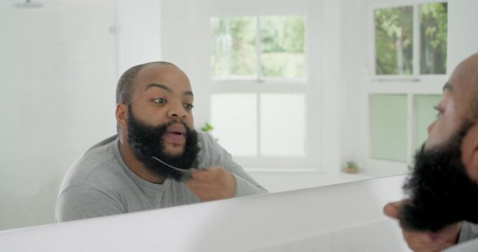 Man wearing gray long-sleeve shirt combing beard and checking angles in bathroom mirror with comb