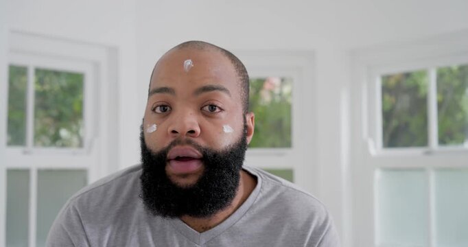 African man in gray shirt at home by windows starting from cheek-cream-dot, dabbing for skincare