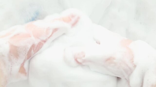Close-up hands gently squeeze and lift dense white foam, slowly kneading airy suds back and forth until the frame fills with soft bubbles in a calming sensory self-care moment.