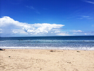 Fototapeta premium Beautiful sandy beach in Bali with surfers riding ocean waves - Tropical coastline under deep blue sky with fluffy white clouds, ideal for summer vacation and travel promos